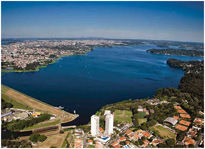 Aerial view of a large blue lake bordered by a city on the left and green hills on the right, with buildings along the shore.