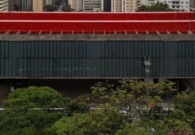 Modern red building with a large glass façade, set behind trees in an urban area.