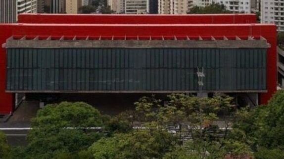 Modern red building with a large glass façade, set behind trees in an urban area.